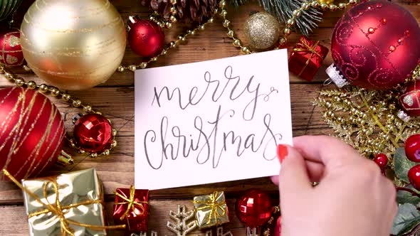 Woman's hand putting a card with the text MERRY CHRISTMAS on a wooden table alt