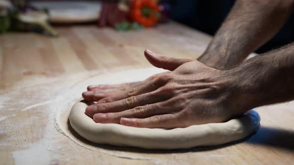 A close-up of a skilled chef's hands pressing and shallowing pizza dough on the wooden counter alt