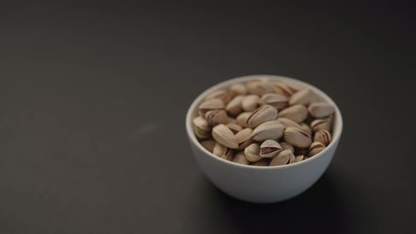Handheld Slow Motion Salted Large Pistachios in White Bowl on Black Paper Background alt