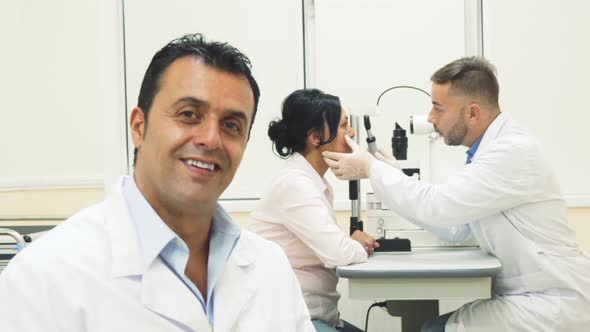 A Smiling Doctor Sits While on the Background Another Doctor Examines the Patient alt