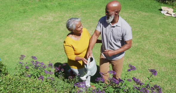 African american senior couple smiling while watering plants together in the garden alt