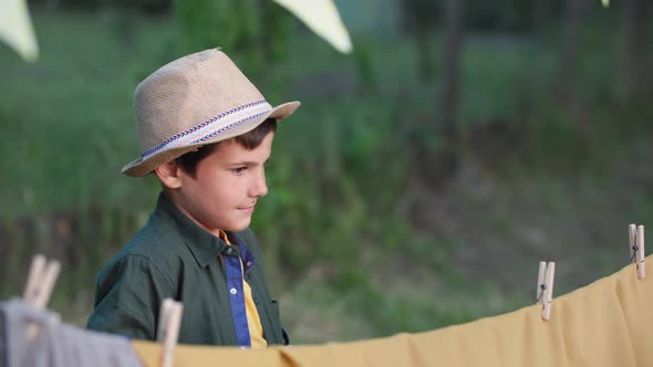 Adorable Boy in a Hat Makes a Wigwam and Clings Clothespins While Relaxing Outdoors on a Summer alt