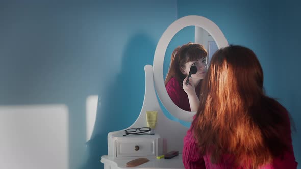 Pretty Woman Sitting in Front of a Mirror at Home and Applying Makeup with a Brush