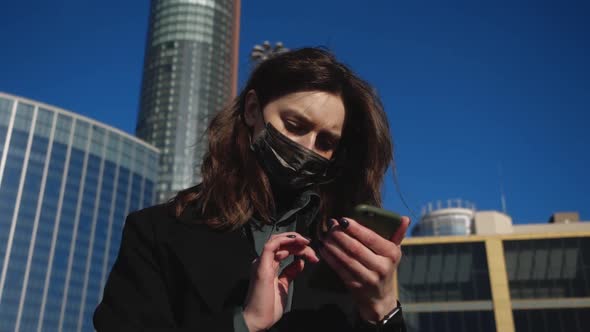 Girl in a Protective Mask Holds a Cell Phone in the Background of the Big City