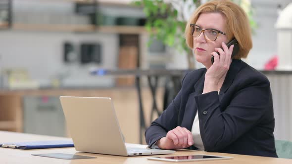 Old Businesswoman with Laptop Talking on Smartphone in Office  alt