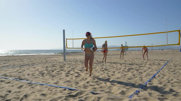 Women players play beach volleyball and a player serves the ball. alt