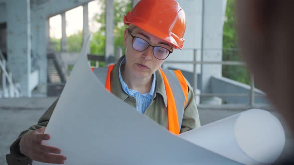 Male Engineer Talking with a Woman in Hard Hat and Safety Vest Holding Blueprint of Building One of