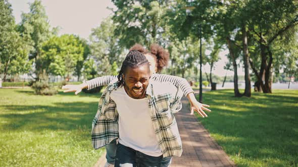 Man Circles His Daughter Sitting on His Piggyback While Walking in a City Park alt