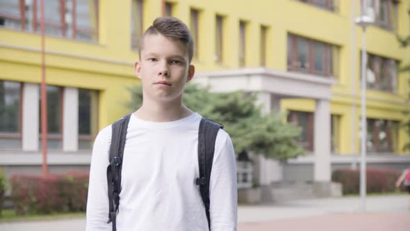 A Caucasian Teenage Boy Looks Seriously at the Camera  a School in the Background