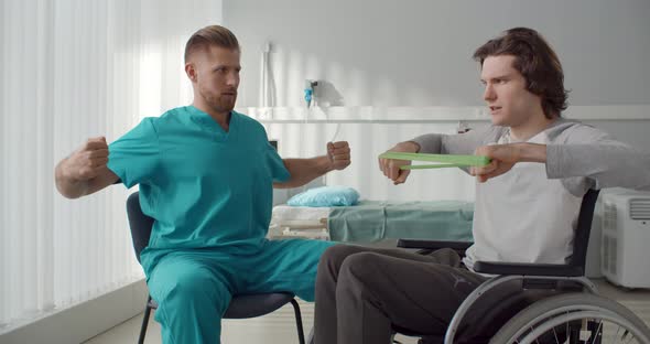 Clinician Helping Young Male Patient in Wheelchair During Physical Exercise with Resistance Band alt