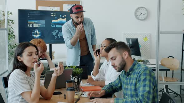 Serious Businessman Making Presentation Talking While Colleagues Playing Games and Putting on Makeup alt