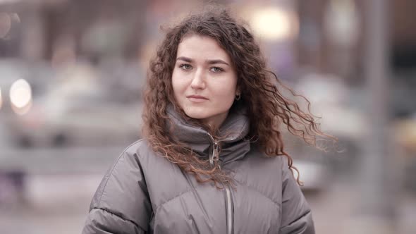 Portrait of a Young Woman with Curly Hair Posing on a City Street on a Cold Day Against the alt