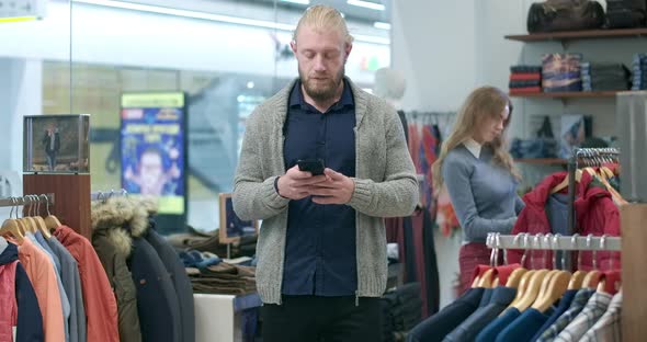Handsome Young Bearded Man Surfing Internet on Smartphone While Woman Choosing Outfit at the alt