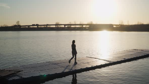 Young athletic girl goes down the pontoon along the lake on the background of beautiful sunset alt