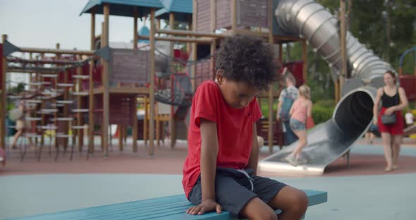 Sad Afroamerican Preschool Boy Sitting Alone on Bench on Playground alt