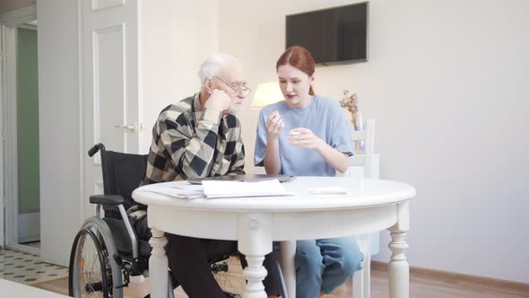 Woman Teaching an Old Man in a Wheelchair How to Use Wireless Headphones alt