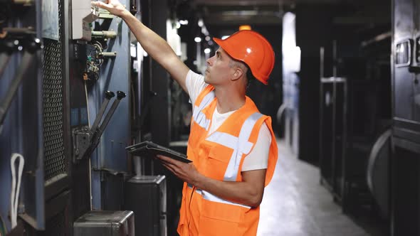 Engineer in Hard Hat and Vest Walks Over to a Rack and Using Digital Tablet Inspect Power System alt