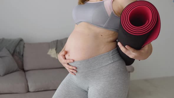 Pregnant Woman in Sportswear Standing with Fitness Mat in Hands at Home in Living Room alt