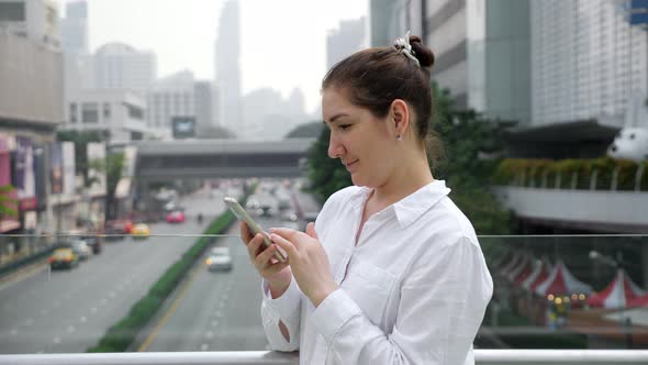 Brunette in White Types on Smartphone on Bridge Over Street alt