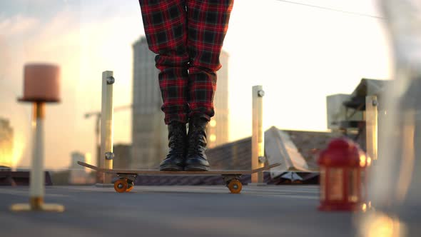 Legs of Unrecognizable Slim Woman in Checkered Pants Standing on Skateboard with Urban Skyline at alt