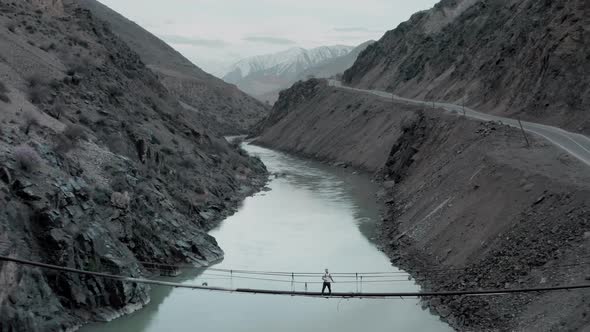 Drone Flies Away From Man Standing on Bridge Over River in Mountain Gorge alt