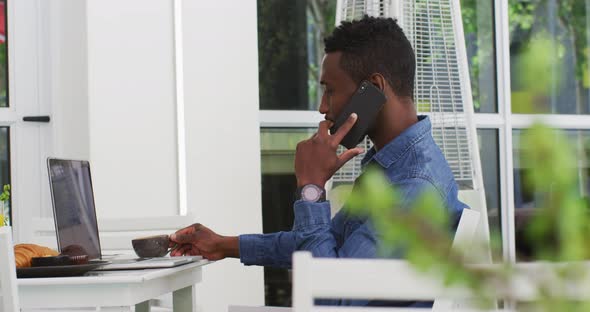 African american businessman using laptop and talking on smartphone in cafe alt