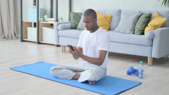 Young African Man Using Smartphone on Yoga Mat at Home alt