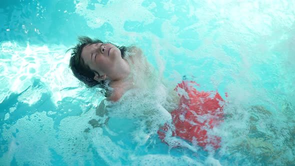 Top View of Playful Cute Boy in Blue Bubbling Water in Swimming Pool Smiling and Looking at Camera alt