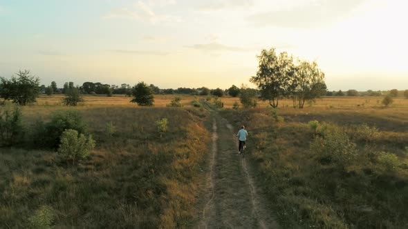 Aerial Footage of Young Woman Riding Bike Through Fields at Summer Sunset alt