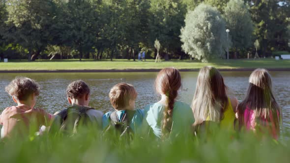 Rear View of Happy Kids Friends Sitting on Grass in Summer Park Near Lake alt