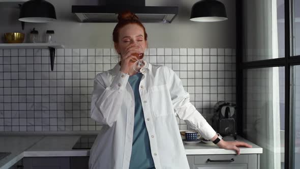 Middle Shot Portrait of Happy Young Woman Drinking Red Wine Standing in Light Kitchen Room at Home alt