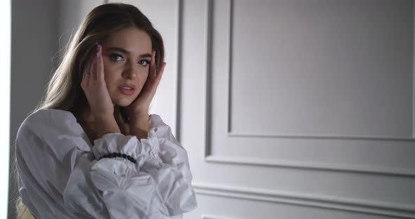 Young Woman Looks at the Camera with a Charming Look, Touching Her Face with Her Hands alt