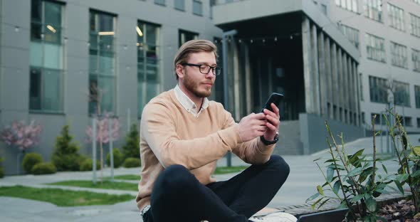 Man in Glasses Using Cellphone while Sitting Crossing Legs on Bench of City Financial District. alt