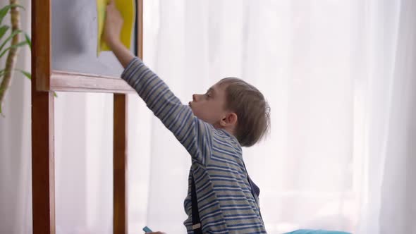 Side View Portrait of Joyful Autistic Schoolboy Cleaning Blackboard with Cloth Indoors in Classroom alt