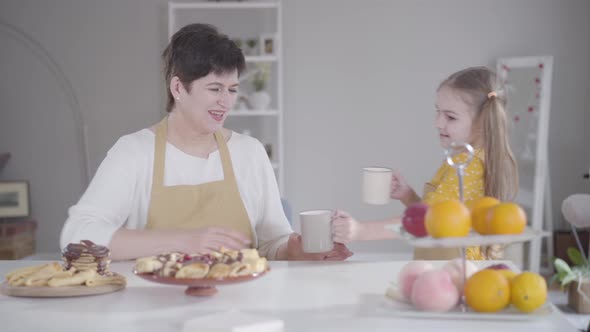 Pretty Little Girl Bringing Cups To the Table with Baked Pancakes. Happy Grandmother and alt