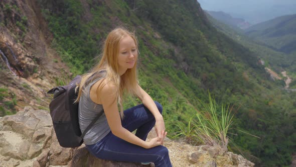 Young Woman-traveler with a Backpack in the Mountains. She Is Looking at the Majestic Valley Down alt