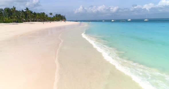 Aerial view of the sea waves, green palm trees, sandy beach, boats in clear blue water