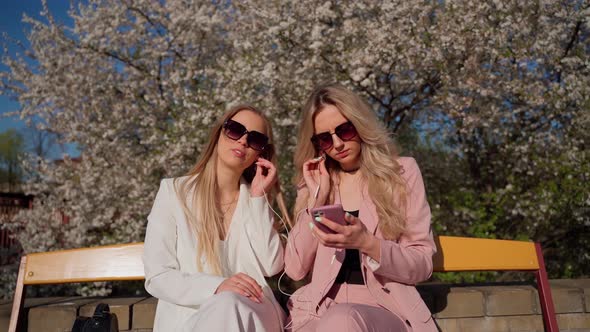 Two females persons looking at smartphone smile and laugh outside. Young adult women in sunglasses alt