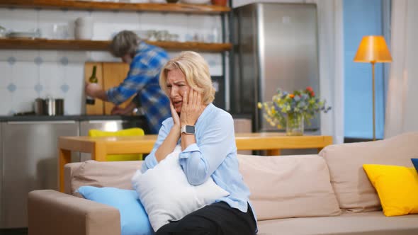 Senior Wife Sitting on Couch and Crying with Husband Drinking on Background alt