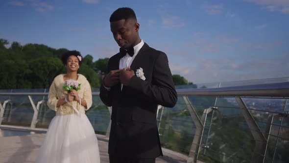 Happy African American Groom Adjusting Wedding Suit and Bow Tie As Smiling Bride in Wedding Dress alt