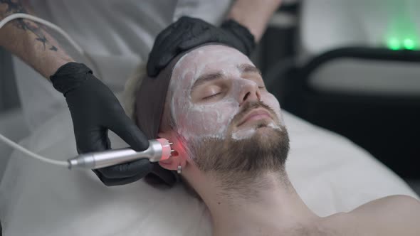 Beautician Doing Red LED Light Peel on Face of Young Handsome Man with Closed Eyes alt