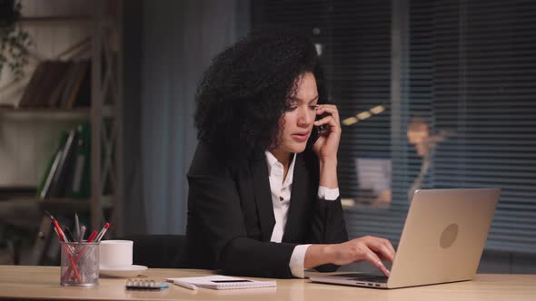 Portrait of Angry African American Woman Typing on Laptop Talking on Smartphone with Indignation alt