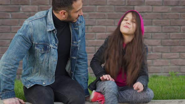 Dad and Daughter Are Sitting, Talking and Chewing Bubble Gum Against the Backdrop of Grass and a alt