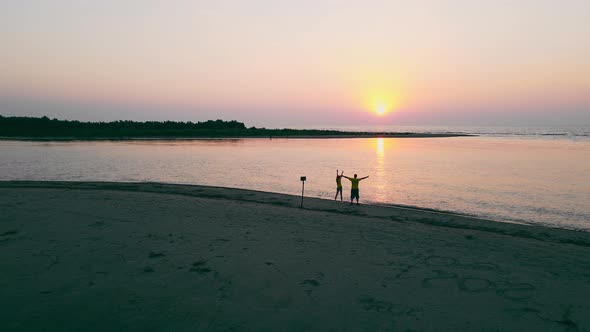 Two Happy People Rejoicing and Raising Their Hands Up By the Sea at Sunrise alt
