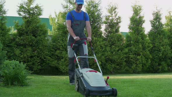 Young Gardener in Overalls Uses a Lawn Mower on the Plot alt