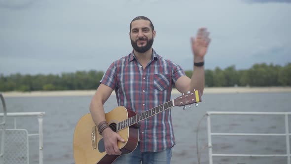 Positive Caucasian Man Standing with Guitar on Embankment and Waving. Portrait of Happy Young alt