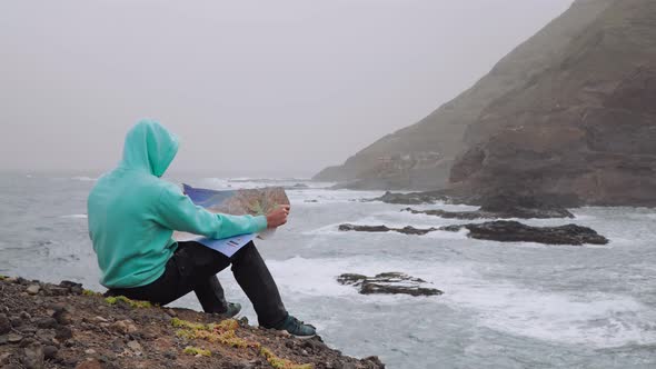 Traveler with Dog Holding Map in Front of Powerful Waves Splashing Into Rocky Coastline alt