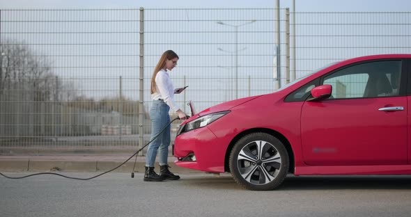 Girl at the Charging Station for Electric Vehicles Inserts a Plug Into the Car Socket