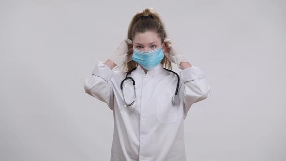 Confident Caucasian Girl in Doctor Gown Putting on Coronavirus Face Mask Crossing Hands Looking at alt