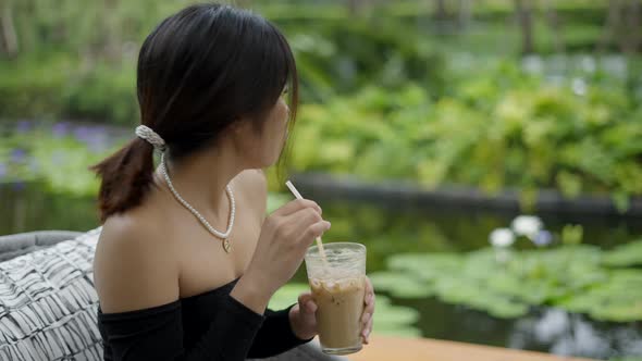 Portrait Shot of Beautiful Young Asian Woman Drinking Cold Coffee in a Green Nature Background alt